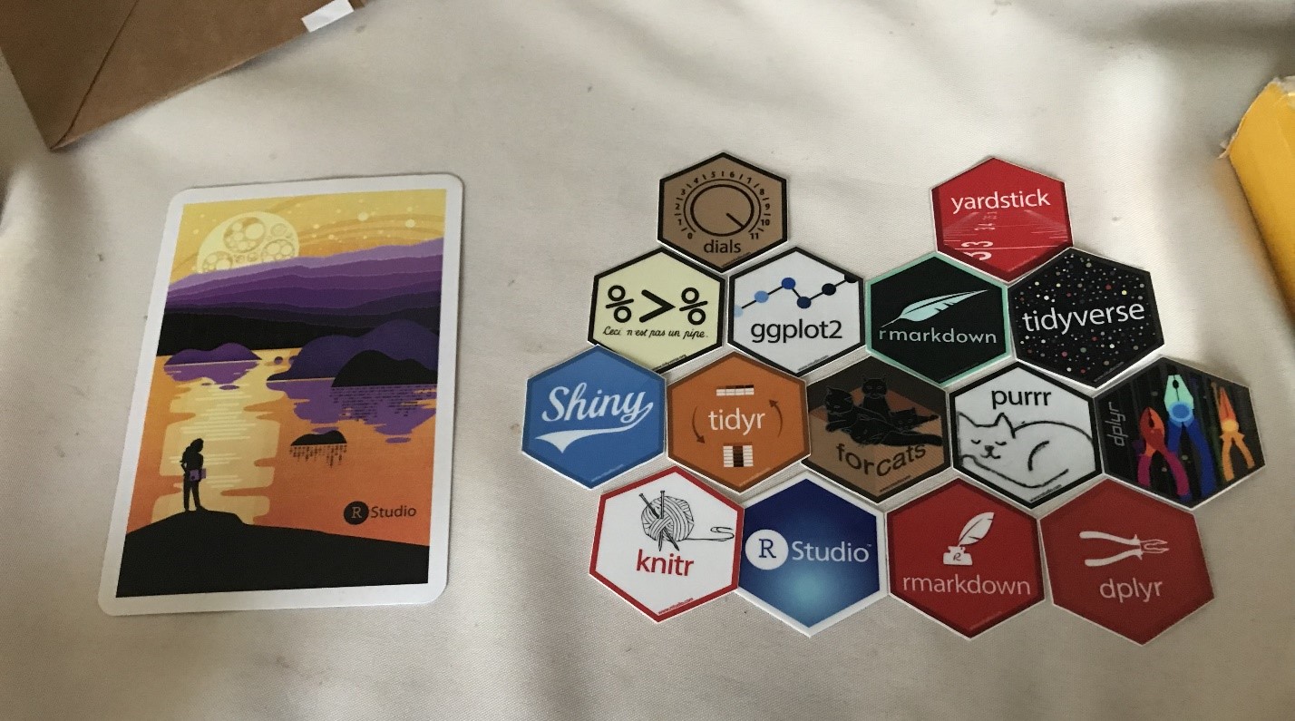 An RStudio postcard and 15 RStudio hex stickers are aligned on a table.